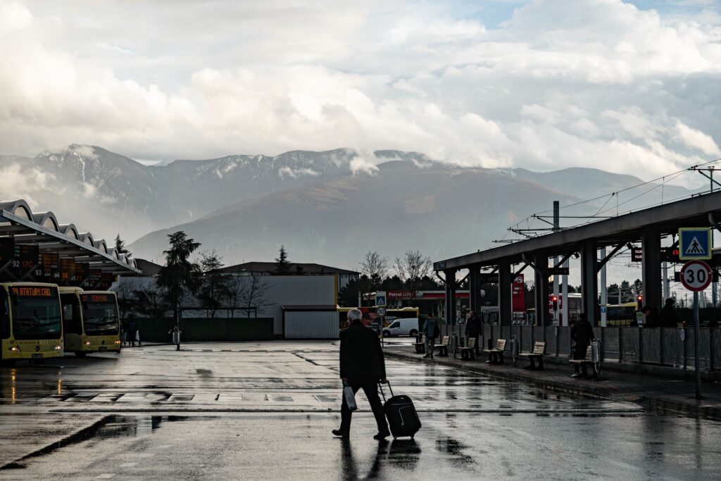 Voyageur avec un sac attendant à un arrêt de bus, montagnes en arrière-plan sous un ciel couvert.