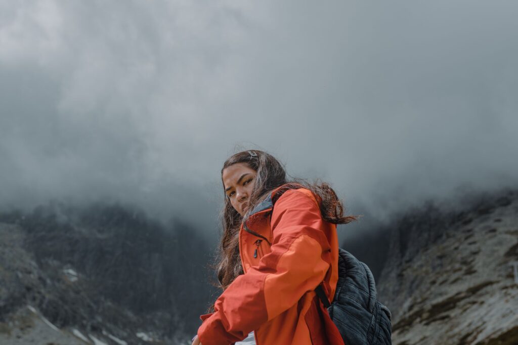Randonneuse en veste coquille orange progressant sur un sentier de montagne noyé dans le brouillard.