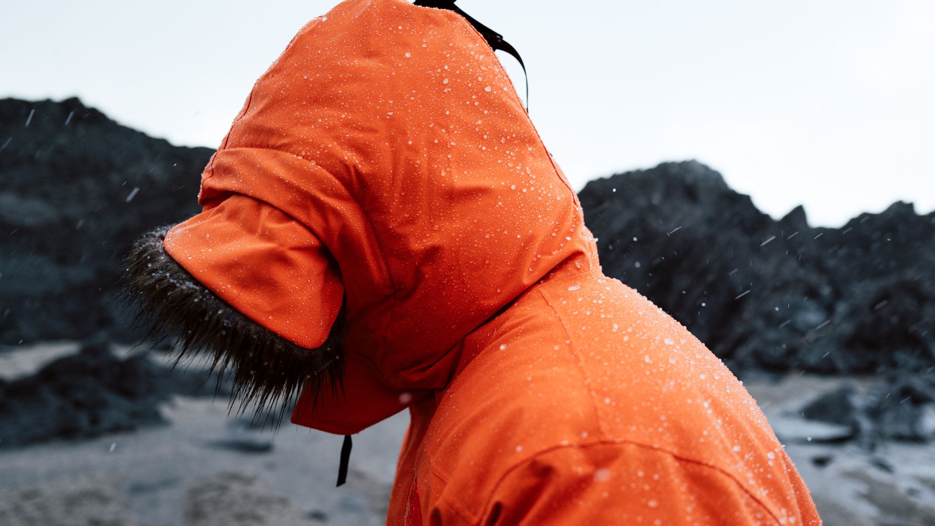 Marcheur en veste orange face à un rivage rocheux sous une pluie hivernale prolongée