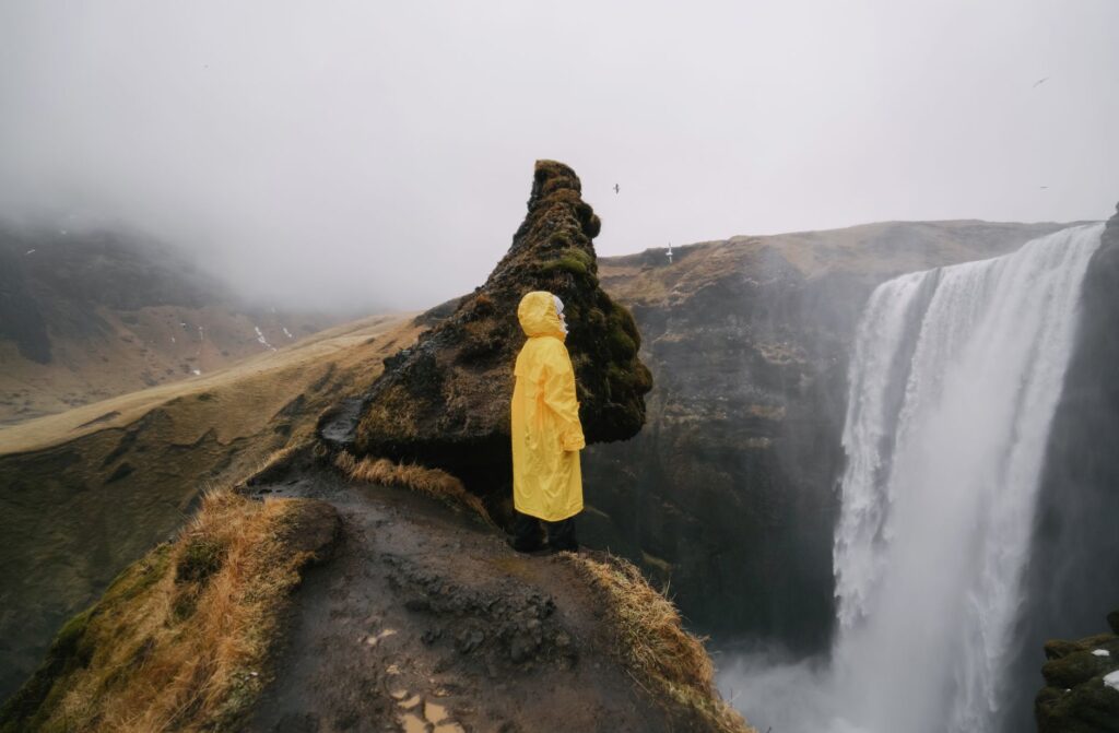 Randonneur en veste de pluie jaune arrêté sur une dalle rocheuse devant une cascade de montagne, par temps de brouillard.