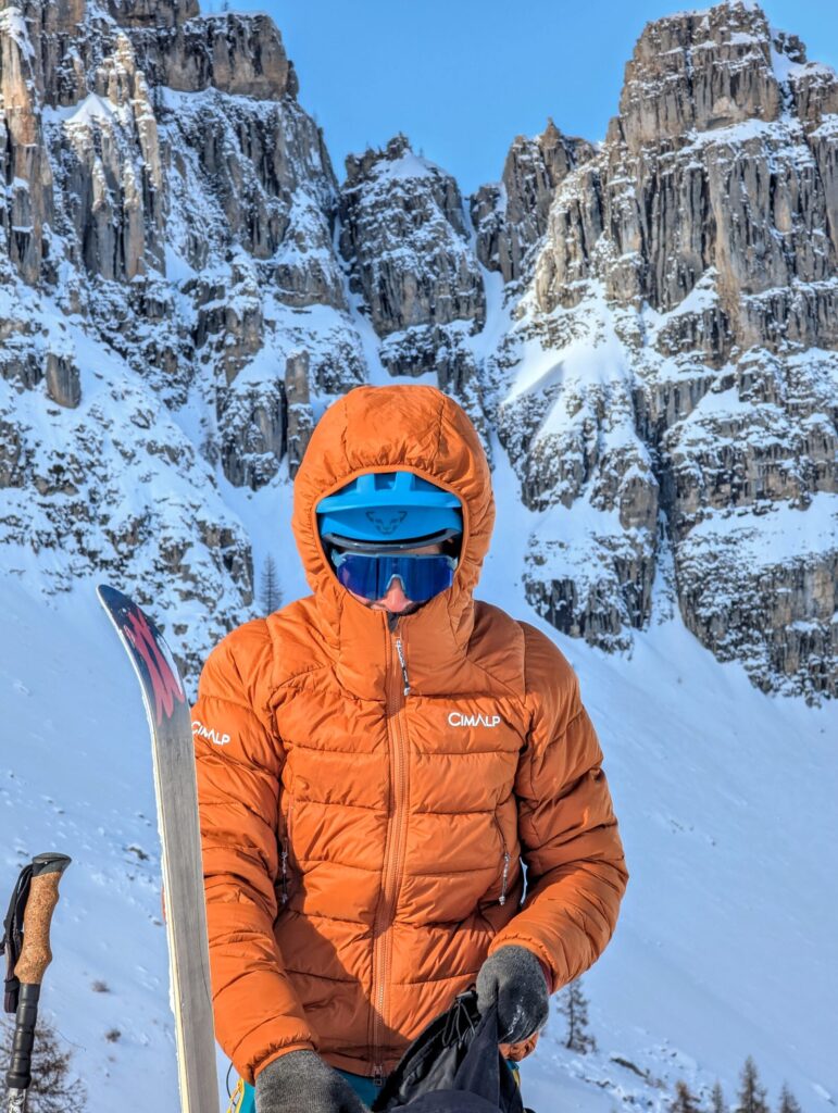 Randonneur en doudoune orange, capuche serrée et masque sur les yeux, debout dans la neige au pied de falaises