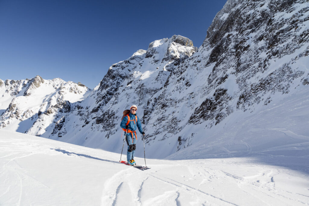 Randonneur à skis au pied de versants raides et rocheux couverts de neige, sous un ciel bleu