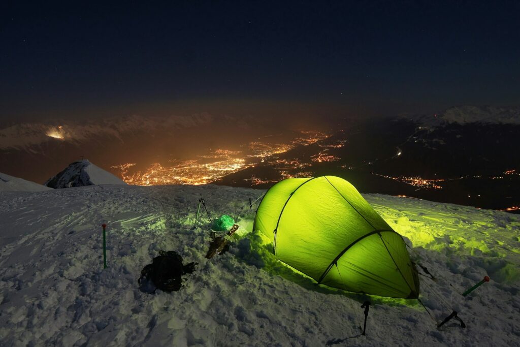 Tente verte éclairée sur une crête enneigée, la nuit, au-dessus des lumières d'une vallée