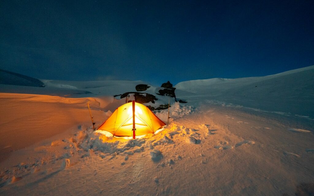 Tente éclairée de l'intérieur, en partie enfoncée dans la neige, sous un ciel de nuit