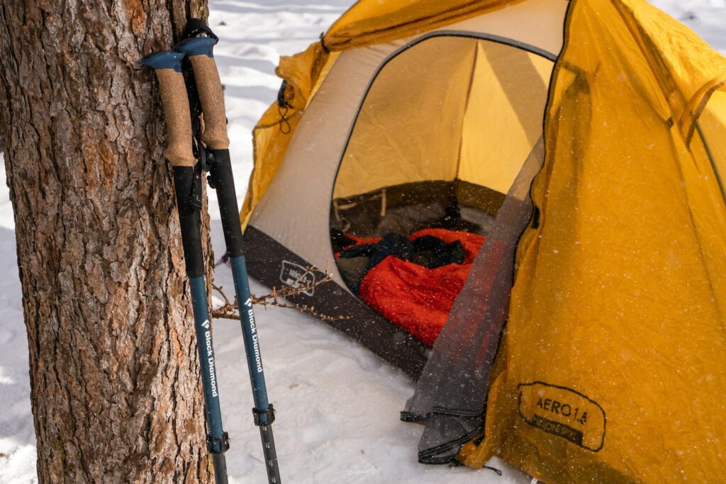 Tente jaune ouverte dans la neige, sac de couchage rouge déplié à l'intérieur, bâtons appuyés contre un arbre